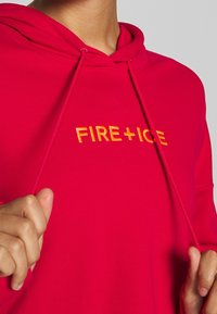 Sudadera roja con cordones elásticos y "FIRE+ICE" bordado en amarillo. Tela suave, corte relajado, detalles de costura visibles.