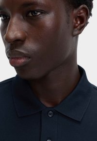 Camisa polo de color marino con una tela texturizada, que presenta un cuello y dos botones negros. Vista en primer plano del cuello y la cara de una persona.