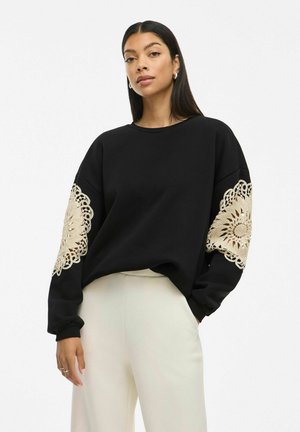 Sweatshirt noir à manches longues avec des détails en dentelle dorée intriquée sur les poignets. Tissu texturé, coupe décontractée, encolure ras du cou.