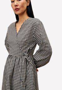 Femme portant une robe à carreaux noire et blanche avec des manches longues, se tenant avec une main derrière le dos contre un fond uni.