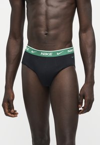 Buste masculin portant un slip Nike noir avec une ceinture verte portant le logo et le texte Nike, debout sur un fond clair uni.