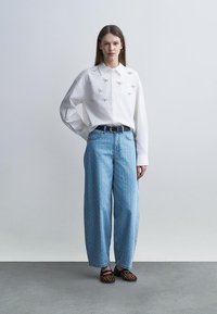 Femme portant une chemise blanche à boutons avec de petits nœuds brodés, un jean bleu clair à jambes larges, une ceinture noire et des ballerines imprimé léopard.