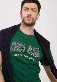 Zielony bawełniany t-shirt z białą i granatową grafiką "CROSS JEANS", z ciemnym swetrem narzuconym na ramiona.