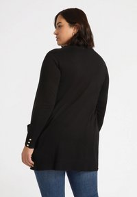 Schwarze Strickjacke mit langen Ärmeln, geripptem Saum und drei goldenen Knöpfen an den Manschetten. Über blauen Jeans getragen, was eine schlanke Silhouette zeigt.