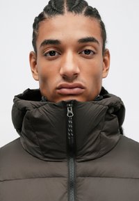 Mørkebrun polstret jakke med høy krage og glidelås, med en glatt tekstur og subtile sømdetaljer.