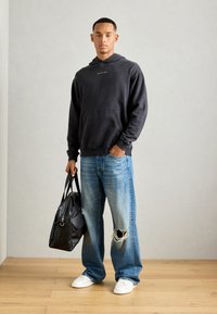 Ung mand iført en mørk hættetrøje, slidte vide jeans, hvide sneakers, holder en sort duffel bag og står på trægulv.