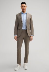Un costume marron clair avec une coupe ajustée, associé à une chemise bleu clair et des baskets blanches. Le costume présente une texture fine et des pantalons slim.