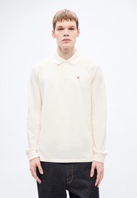 Garām piedurknēm polo krekls gaiši bēšā kokvilnā ar apkakli un trim pogām. Kreisajā krūtīs ir mazs sarkans logotips.