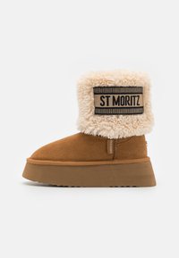 Stivaletto alla caviglia in suede color tan con un morbido collo in pelliccia sintetica color crema, dotato di un'etichetta "ST MORITZ" nera e beige e di una suola texturizzata.