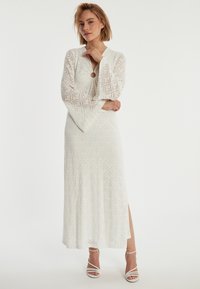 Witte maxi-jurk met lange klokmouwen, voorzien van een diamanten patroon, een ronde uitsnijding bij de hals en een zij-split. Gecombineerd met witte sleehakken.