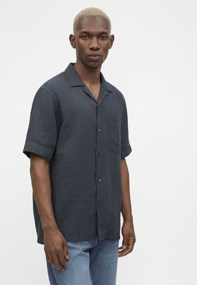 Άνδρας με κοντά ξανθά μαλλιά που φοράει σκούρο γκρι πουκάμισο με κοντά μανίκια και ανοιχτό μπλε τζιν, στέκεται μπροστά από ένα απλό φόντο.