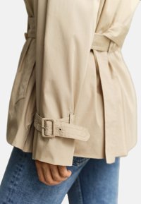 Μανίκι μπεζ μπουφάν με ιμάντα και αγκράφα, ρυθμιζόμενο, φορεμένο πάνω από μπλε τζιν σε χέρι και μέση ατόμου.