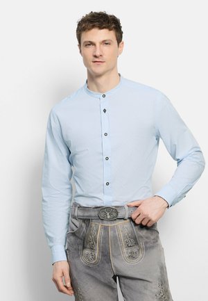 Man draagt een lichtblauw overhemd met knopen en traditionele grijze geborduurde lederhosen, en stelt de gesp van de riem met zijn rechterhand af.