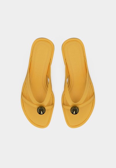 Κίτρινα σανδάλια τύπου thong με επενδυμένες σόλες και στρογγυλές σκοτεινές διακοσμήσεις στα μπροστινά λουριά, εμφανίζονται από πάνω σε λευκό φόντο.