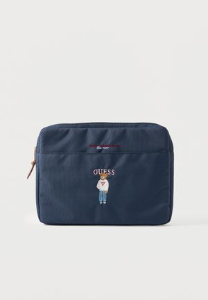 Pochette bleu marine avec zip, ours brodé portant un pull et un jean, et texte « GUESS » au centre devant.