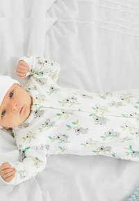 Valkoinen vetoketjullinen haalari, jossa on koalan kuva harmailla ja vihreillä yksityiskohdilla, valmistettu pehmeästä kankaasta, ja sitä on täydennetty sopivalla valkoisella myssyllä.