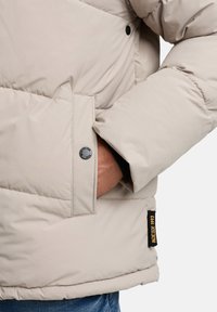 Lys beige pufferjakke med en glat overflade, store quiltede sektioner, trykknaplukning, sidelomme og et sort mærke med teksten "1993."