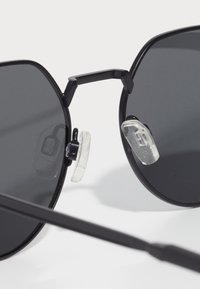 Ochelari de soare negri cu o formă unghiulară. Lentile întunecate, cadru din metal ușor și pernițe nazale din silicon clar pentru confort și stabilitate.