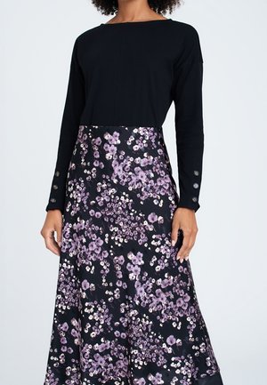 Mujer que lleva una blusa negra de manga larga y una falda negra con un estampado floral en púrpura y blanco, de pie frente a un fondo liso.