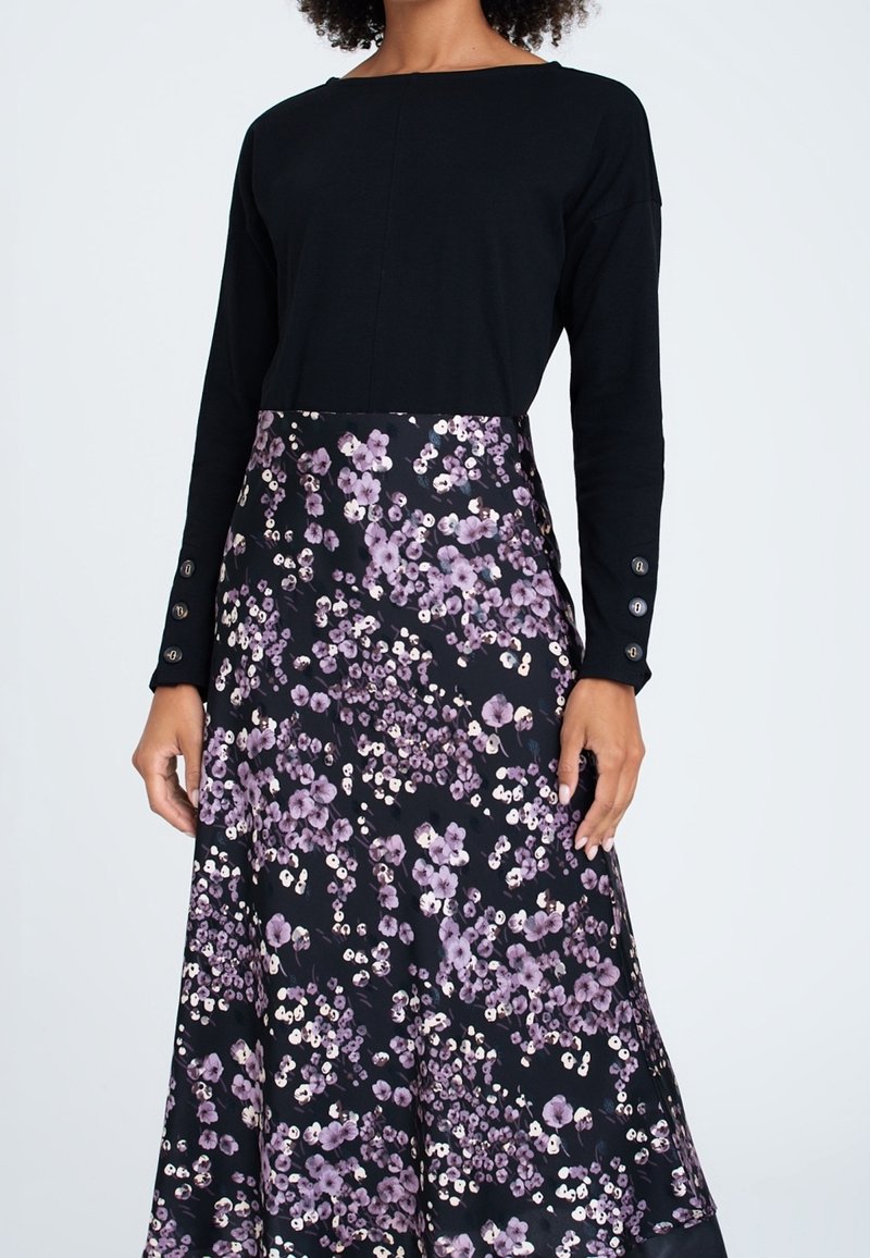 Femme portant un haut noir à manches longues et une jupe noire avec un motif floral violet et blanc, debout devant un fond uni.