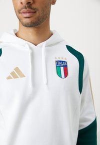 Λευκό φούτερ με πράσινες λεπτομέρειες, που φέρει το εθνικό έμβλημα ποδοσφαίρου της Ιταλίας και το χρυσό λογότυπο της adidas, σε συντονισμένα παστέλ υφάσματα.