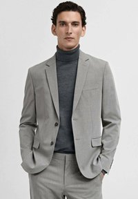 Costume gris ajusté avec un blazer à simple boutonnage, des revers crantés et un pantalon assorti. Porté sur un pull col roulé gris foncé.