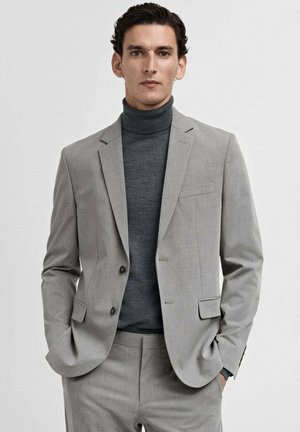 Grau maßgeschneiderter Anzug mit einem einreihigen Blazer, mit Revers und passender Hose. Trägt ein dunkelgraues Rollkragenpullover darunter.
