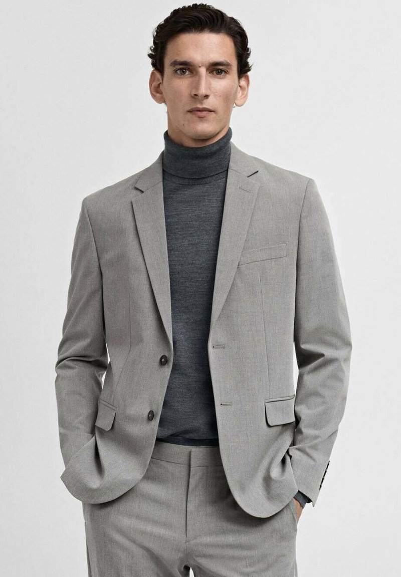 Costume gris ajusté avec un blazer à simple boutonnage, des revers crantés et un pantalon assorti. Porté sur un pull col roulé gris foncé.