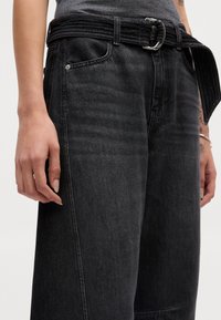 Črne denim hlače imajo širok kroj, pleten pas in rahlo obrabo. Vključujejo sprednje žepke in detajle šivanja vzdolž šivov.