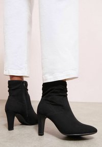 Botines negros con una parte superior arrugada, punta afilada y tacón de aguja de 3 pulgadas, fabricados con un tejido suave y con una cremallera en el lateral.