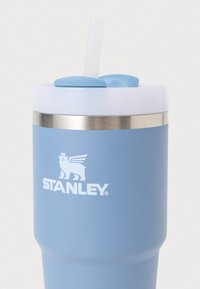 Modrý znovu použitelný pohár s nerezovým páskem, bílým víčkem a průhlednou brčkem. Emblém s korunovaným medvědem a textem "STANLEY" na těle.