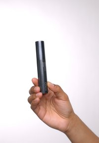 Zwarte cilindrische mascara tube met een matte textuur, voorzien van zilveren letters en een stevige dop, vastgehouden tussen de vingers.
