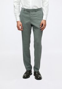 Pelēki teksturēti uzvalka bikses ar pielāgotu piegriezumu un gludu priekšpusi, ar jostu cilpiņām un sānu kabatām, saskaņotas ar melnām kurpēm.