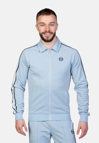 Helle blaue Zip-Jacke mit Kragen, ausgestattet mit schwarzen Seitenstreifen und Logo. Slim Fit mit gerippten Bündchen und Taschen.