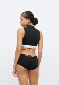 Μαύρο κοντό τοπ με ψηλό λαιμόκοψη και λευκή ζώνη. Μαύρο σλιπ με το λευκό λογότυπο Calvin Klein στη ζώνη μέσης. Ομαλή υφή από βαμβάκι.