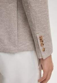 Blazer marron clair texturé avec un petit motif, doté de quatre boutons ronds beige au niveau des poignets et d'une coupe nette et ajustée.