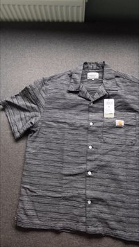 Camicia a maniche corte con bottoni a righe alternate bianche e nere, caratterizzata da una tasca sul petto e un'etichetta che indica la taglia XL.