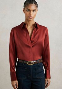 Cămașă roșie din satin cu guler ascuțit, are mâneci lungi și închidere cu nasturi, purtată cu blugi negri din denim și o curea maro cu o cataramă aurie.