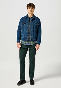 Jeansjack in diepblauw, met knopen aan de voorkant en twee borstzakken over een geruit overhemd in groen, blauw en wit, gecombineerd met groene broek.