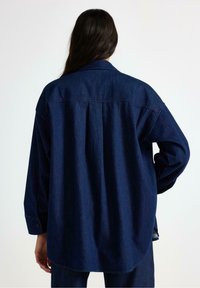 Mörkblå denimskjorta med krage, långa ärmar och rundad nederkant. Har en central veckning och knäppte manschetter. Mjuk textur.