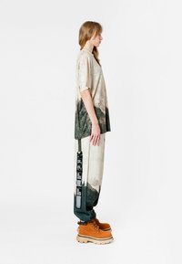 Camisa holgada de manga corta con un estampado texturizado de paisaje en beige y verde; combinada con pantalones amplios estampados y botas marrones.