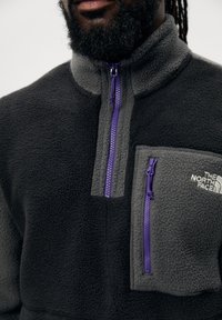 Fleece-pullover i sort og grå med høj krave. Har en lilla lynlås og en brystlomme med brandlogo. Blød tekstur.
