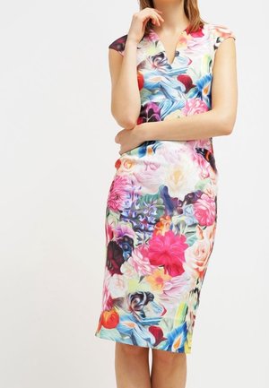 Vrouw draagt een mouwloze, knielange jurk met een kleurrijk, abstract bloemenpatroon op een lichte achtergrond.