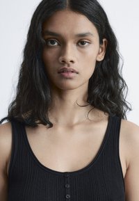 Black ribbed tank top with a scoop neckline, featuring a button detail down the front. Made of soft fabric with a textured finish.