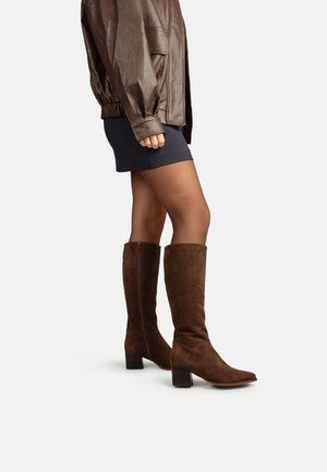 Vrouw die bruine suède kniehoge laarzen met blokhakken, een marineblauwe minirok en een bruine leren jas draagt, staand zijwaarts.