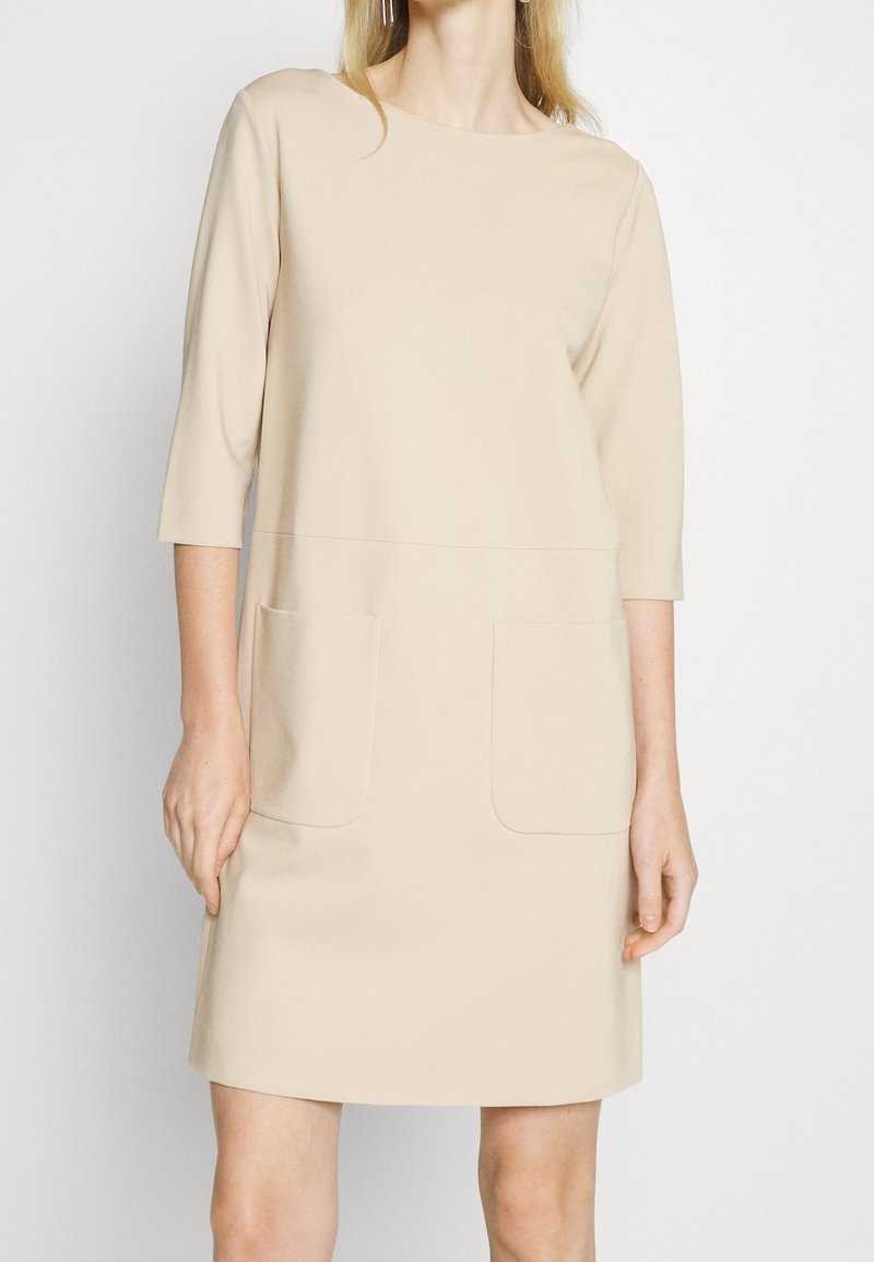 Femme portant une robe beige longueur genoux avec des manches trois-quarts et deux poches avant sur un fond uni.