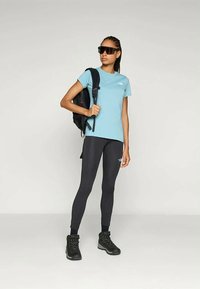 Világoskék, testhezálló póló, fekete leggings és strapabíró fekete túracipő. Hátizsák egy vállon, napszemüveg, semleges háttér előtt.