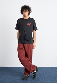 T-shirt preto com gráfico vermelho, calças largas vermelhas, bolsa tiracolo preta, sapatilhas pretas com detalhes brancos, em pé sobre um carpete azul.