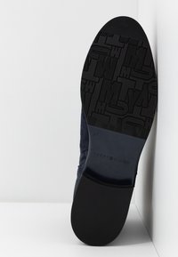 Semelle d'une chaussure foncée montrant un motif texturé avec des lettres et la marque "Tommy Hilfiger" près du talon sur un fond blanc.