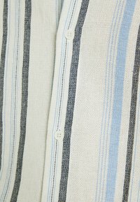 Camisa a rayas con una tela texturizada en blanco, azul claro y negro. Cierre de botones con dos botones visibles. Patrón vertical.